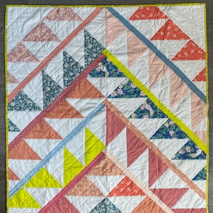 May include: A square quilt with a white background and a geometric pattern of triangles in various colours, including coral, pink, teal, and yellow. The quilt is bordered with a bright yellow binding and features diagonal lines.