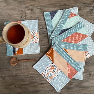 May include: Handmade fabric coasters with patchwork designs in blue, orange, and beige. The coasters feature floral, dotted, and striped patterns. A mug of tea and a wooden spoon are also visible. The coasters are approximately 10 cm square.