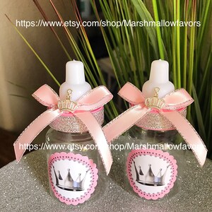 May include: Two clear plastic baby bottles decorated with pink bows, glitter, and crown stickers. The bottles have white tops and are embellished with small gold crowns. The text "https://www.etsy.com/shop/Marshmallowfavors" is visible.