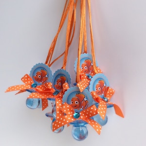 May include: Set of six blue and orange pacifier favors with orange and white polka dot bows and orange ribbon. Each pacifier has a blue and white circular tag with a cartoon image of a clownfish.