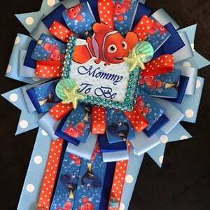 May include: A blue and orange baby shower sash with a Finding Nemo theme. The sash has white polka dots and features a picture of Nemo, the clownfish, with the text "Mommy To Be".