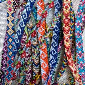 May include: A collection of multiple woven friendship bracelets featuring intricate geometric patterns, zigzags, and small strawberry motifs. The bracelets are crafted from colorful embroidery thread in shades of blue, orange, red, pink, and yellow.