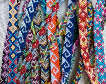 Può includere: Una collezione di diversi braccialetti dell'amicizia intrecciati con complessi motivi geometrici, zig-zag e piccoli motivi a fragola. I braccialetti sono realizzati con filo da ricamo colorato nelle tonalità del blu, arancione, rosso, rosa e giallo.
