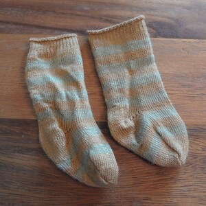 May include: A pair of ankle-length knit socks featuring horizontal stripes in alternating shades of light tan and soft sage green. The socks have a simple, unadorned cuff at the top and a standard heel construction, crafted with a fine, uniform knit texture.