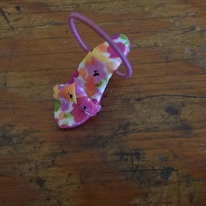 May include: A miniature high-heeled shoe with a floral pattern in pink, orange, and white, accented with a purple strap. The shoe has a small bow detail on the front.