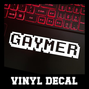 May include: White pixelated "GAYMER" vinyl decal on a black surface, with the text "VINYL DECAL" below. The background features a laptop keyboard with red illuminated keys.