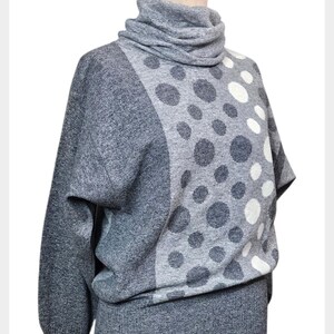 May include: A grey sweater with a cowl neck and a distinctive polka dot design. The sweater has a mix of dark grey and white polka dots on a lighter grey background. The sleeves are a solid grey colour.