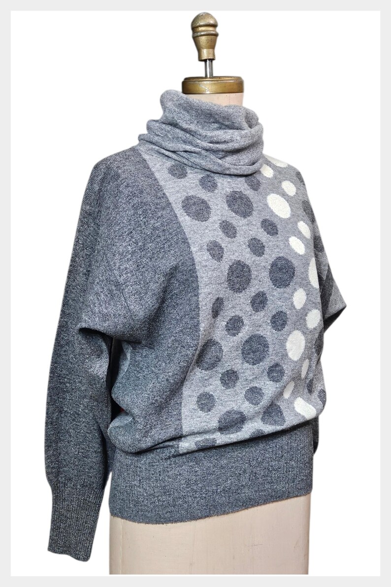 May include: A grey sweater with a cowl neck and a distinctive polka dot design. The sweater has a mix of dark grey and white polka dots on a lighter grey background. The sleeves are a solid grey colour.