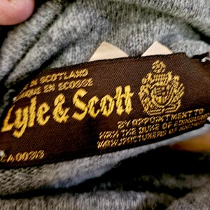 May include: A close-up shot of a brown label with gold lettering, reading "Lyle & Scott", with a crest. The label indicates the item was made in Scotland. The background is a grey knitted fabric.