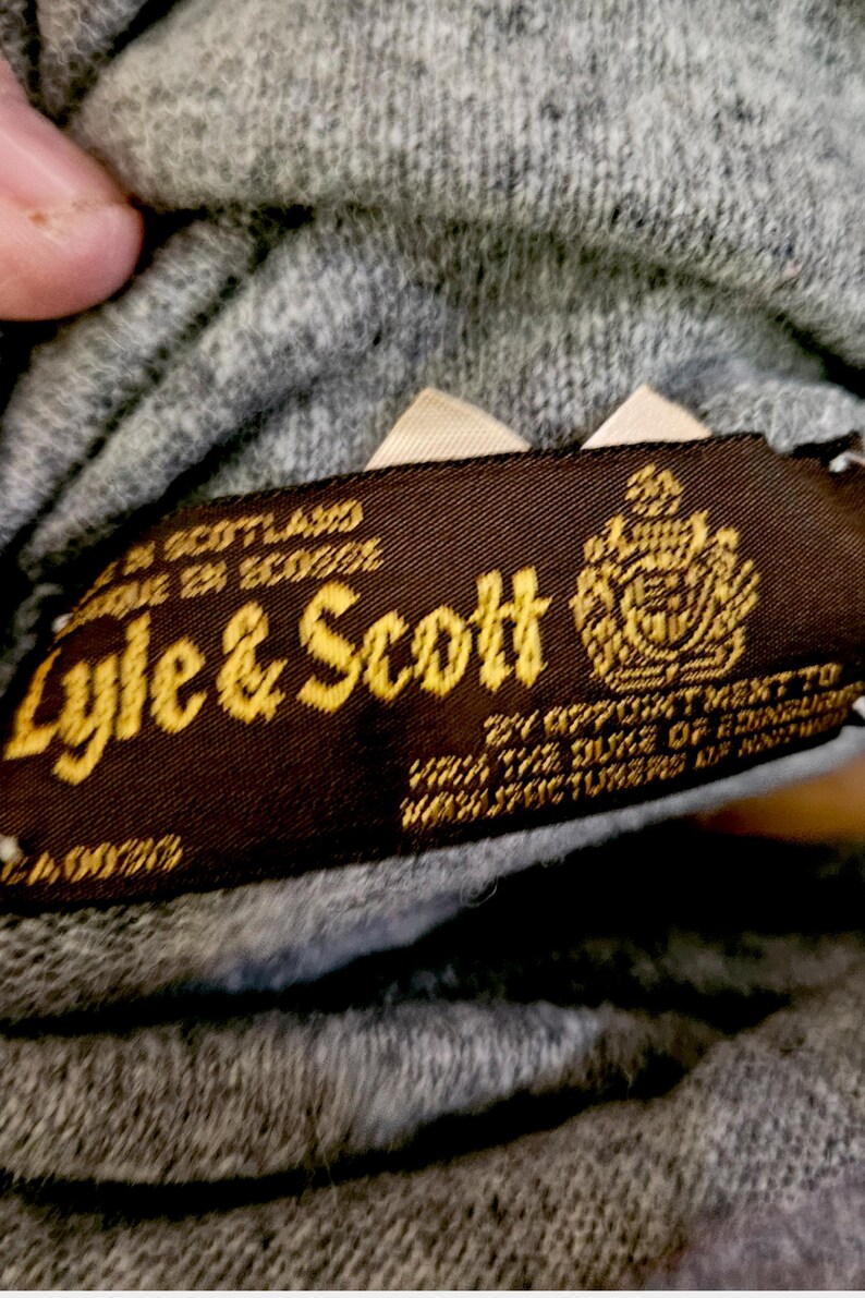 May include: A close-up shot of a brown label with gold lettering, reading "Lyle & Scott", with a crest. The label indicates the item was made in Scotland. The background is a grey knitted fabric.