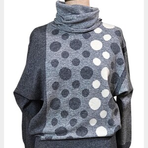 May include: A grey turtleneck jumper with a distinctive polka dot design. The jumper has a high, folded collar and long sleeves. The polka dots are in shades of grey and white, contrasting against the solid grey background. The jumper is displayed on a mannequin.