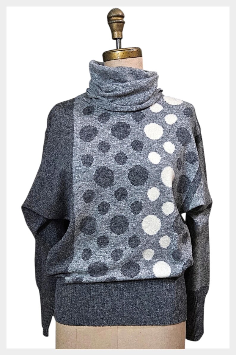 May include: A grey turtleneck jumper with a distinctive polka dot design. The jumper has a high, folded collar and long sleeves. The polka dots are in shades of grey and white, contrasting against the solid grey background. The jumper is displayed on a mannequin.