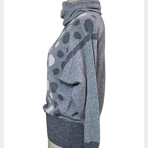 May include: A grey sweater with a high turtleneck collar and long sleeves. The sweater has a pattern of large, irregular white and dark grey polka dots. The cuffs and hem are a darker shade of grey.
