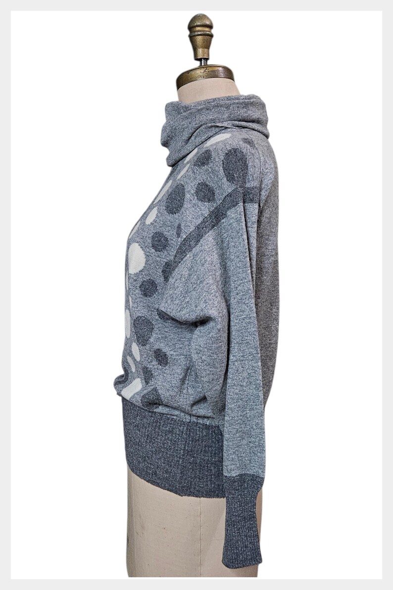 May include: A grey sweater with a high turtleneck collar and long sleeves. The sweater has a pattern of large, irregular white and dark grey polka dots. The cuffs and hem are a darker shade of grey.