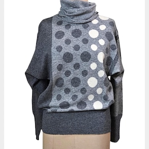 May include: A grey turtleneck jumper featuring a pattern of grey and white polka dots of different sizes. The jumper has long sleeves and a ribbed hem. The garment is displayed on a mannequin.