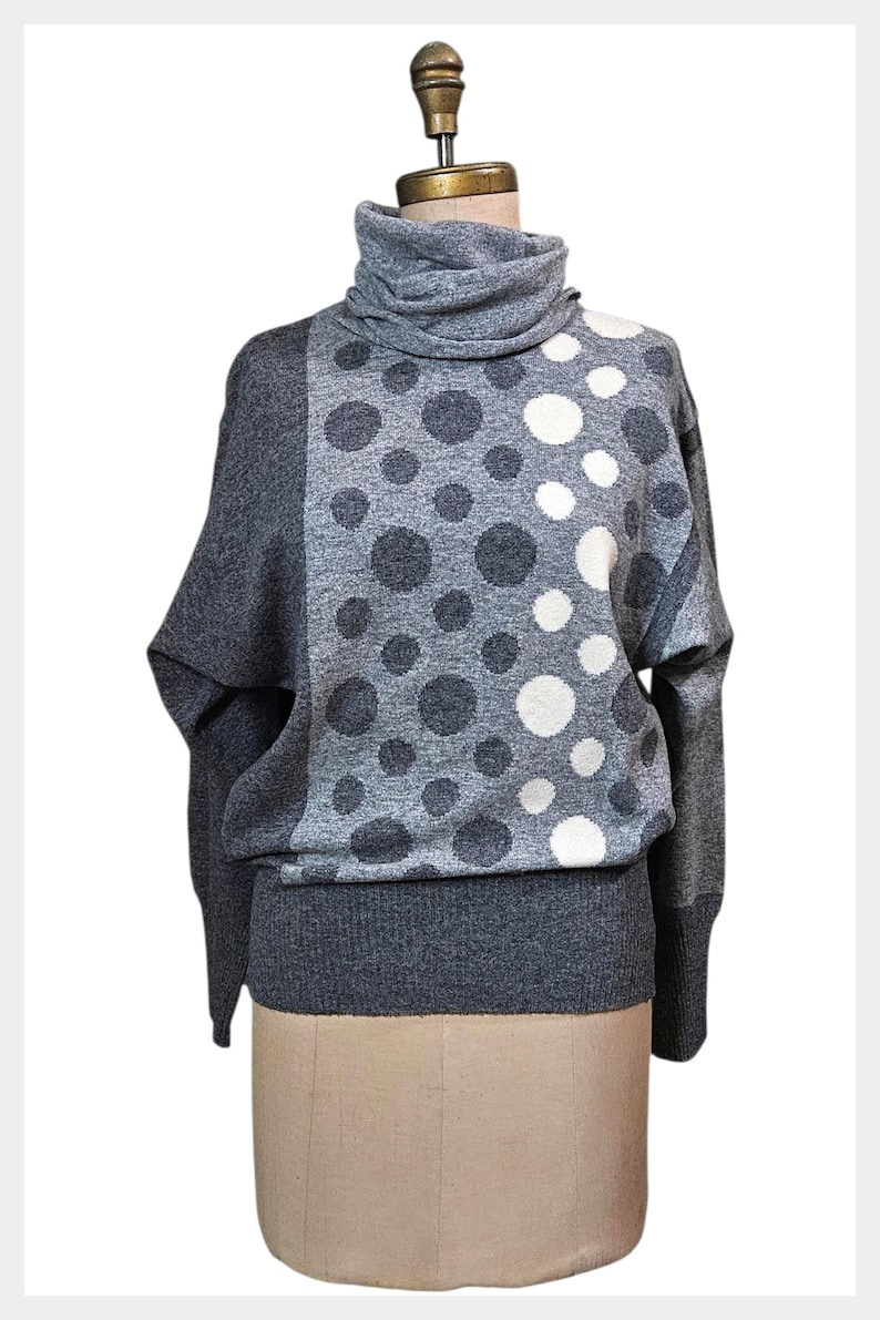 May include: A grey turtleneck jumper featuring a pattern of grey and white polka dots of different sizes. The jumper has long sleeves and a ribbed hem. The garment is displayed on a mannequin.