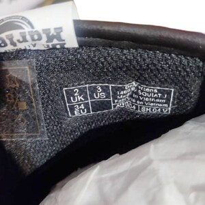 May include: Close-up of the inside of a black leather shoe with a size tag that reads "2 UK, 3 US, 34 EU". The tag also includes the text "Dr. Martens 1460 8-eye SQUIAT J Made in Vietnam Fabriqu&eacute; en Vietnam AV004 SH 04 V."