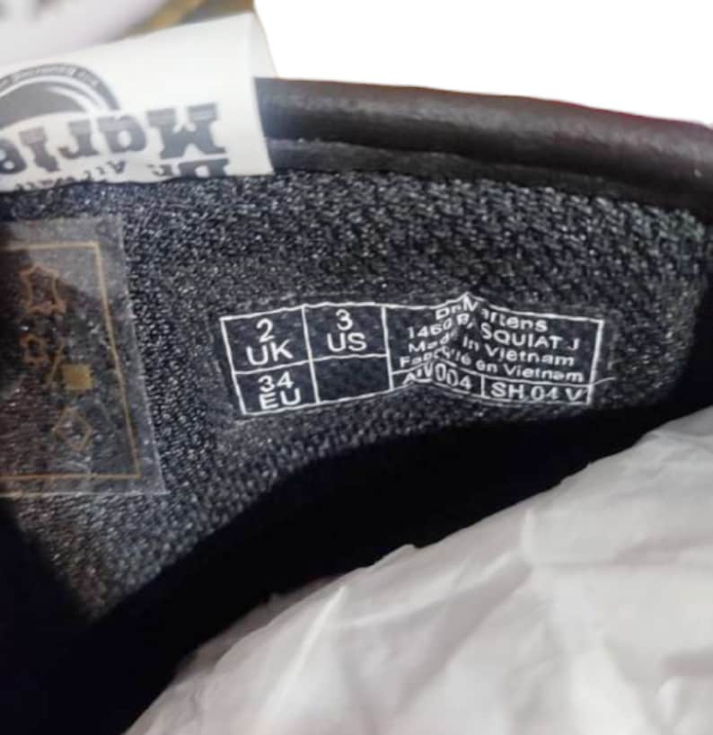 May include: Close-up of the inside of a black leather shoe with a size tag that reads "2 UK, 3 US, 34 EU". The tag also includes the text "Dr. Martens 1460 8-eye SQUIAT J Made in Vietnam Fabriqu&eacute; en Vietnam AV004 SH 04 V."