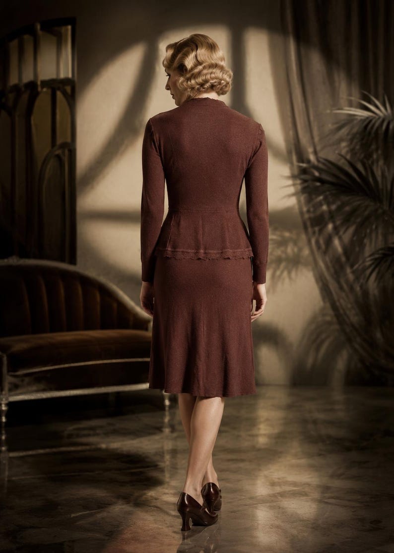 Op de afbeelding: Een donkerbruine tweedelige set bestaande uit een top met lange mouwen en een peplum taille en een bijpassende rok tot op de knie. De top heeft een hoge halslijn en een subtiele kanten rand langs de peplum, gemaakt van fijn breiwerk.