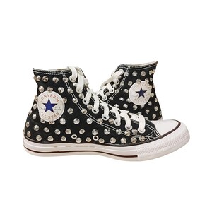 Puede incluir: Zapatillas altas negras adornadas con tachuelas plateadas. Cuentan con cordones blancos, puntera de goma blanca y suela blanca con una franja roja. El logotipo de Converse All Star es visible en el lateral.