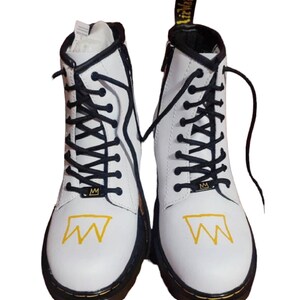 May include: White leather ankle boots with black laces and a yellow crown graphic. The boots have a black rubber sole.