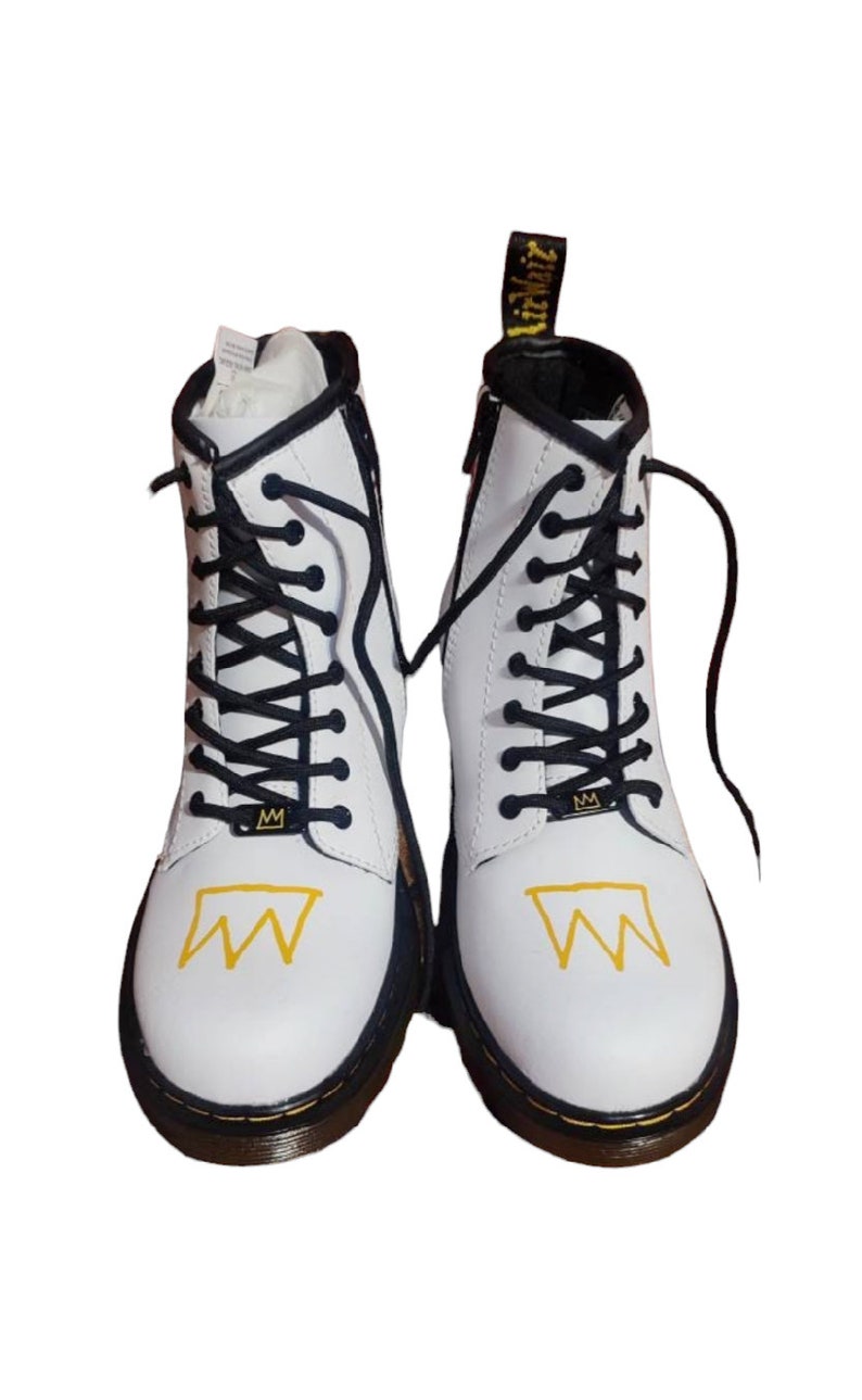 May include: White leather ankle boots with black laces and a yellow crown graphic. The boots have a black rubber sole.