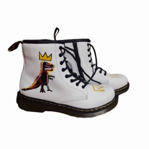 May include: White leather Dr. Martens boots with black laces and a yellow crown and dinosaur graphic.