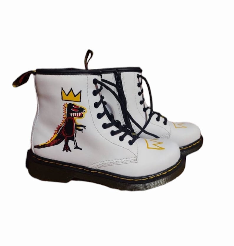 May include: White leather Dr. Martens boots with black laces and a yellow crown and dinosaur graphic.