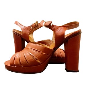 Puede incluir: Un par de sandalias de plataforma de cuero marrón con un tacón grueso. Las sandalias tienen un diseño cruzado en la parte delantera y una correa de tobillo.