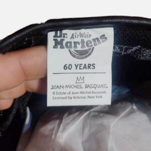 May include: A black leather Dr. Martens shoe with a white label that says "Dr. AirWair Martens 60 YEARS" and "JEAN-MICHEL BASQUIAT&trade; Estate of Jean-Michel Basquiat. Licensed by Artestar, New York"
