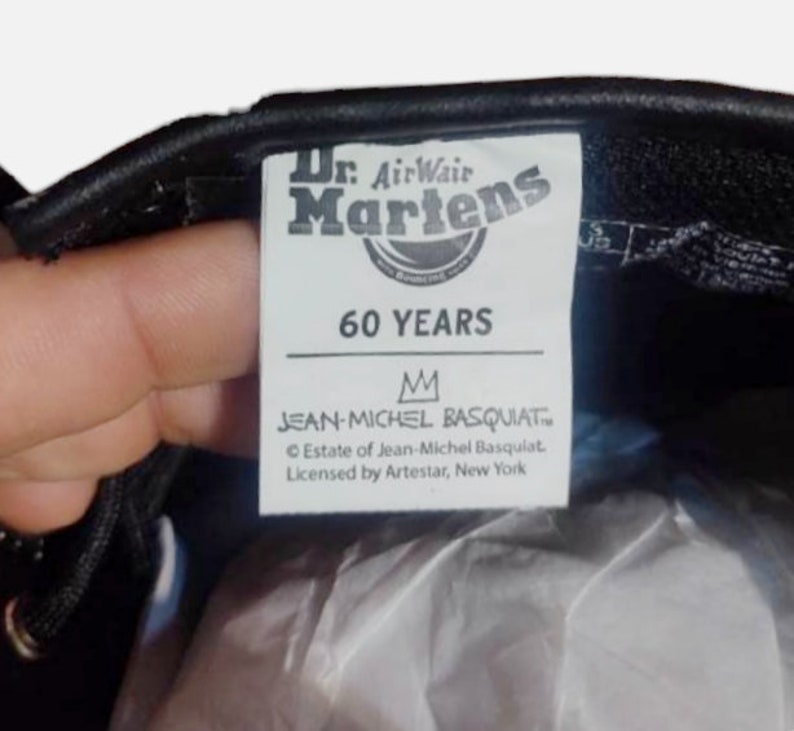 May include: A black leather Dr. Martens shoe with a white label that says "Dr. AirWair Martens 60 YEARS" and "JEAN-MICHEL BASQUIAT&trade; Estate of Jean-Michel Basquiat. Licensed by Artestar, New York"
