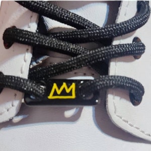 May include: White leather shoe with black laces and a black metal lace charm with a yellow crown design.