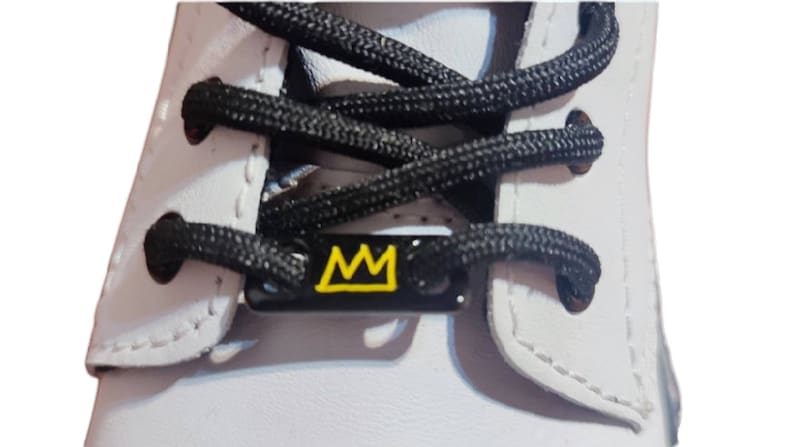 May include: White leather shoe with black laces and a black metal lace charm with a yellow crown design.