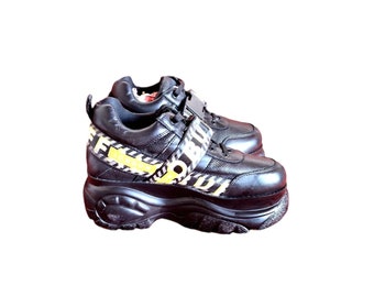 Puede incluir: Zapatillas negras de plataforma con una correa a rayas amarilla y negra y la palabra "FORBIDDEN" impresa en la correa.