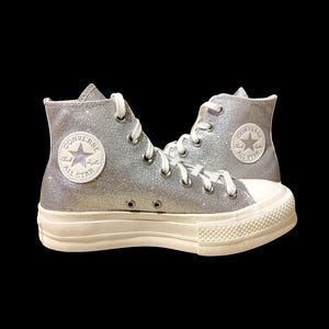 Puede incluir: Zapatillas altas plateadas con purpurina, cordones blancos y suela de goma blanca. Las zapatillas presentan el icónico logotipo de Converse All Star en el lateral. Una elección de calzado elegante y llamativa.
