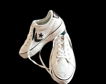 May include: White canvas sneakers with black trim and a black star logo on the side. The shoes have white laces and a black heel patch. The soles are white with a black stripe.