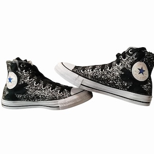 Puede incluir: Par de zapatillas altas Converse All Star negras con detalles de purpurina plateada. Las zapatillas tienen suelas y punteras de goma blanca, cordones negros y el parche del logo Converse All Star con una estrella azul.
