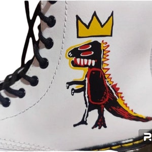 May include: White leather boots with black laces and a yellow crown and red, black, and yellow dinosaur graphic.