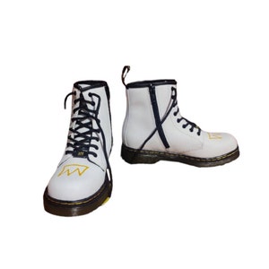 May include: White leather ankle boots with black laces and a yellow crown design. The boots have a black zipper on the side and a black and yellow sole.