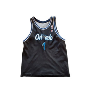 Può includere: Maglia da basket nera con bordini blu e bianchi. La maglia ha "Orlando" scritto sul davanti con una stella e il numero "1" sotto.