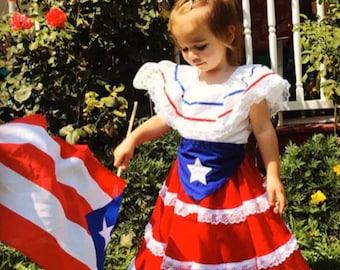 May include: A young child wearing a traditional Puerto Rican dress, holding a Puerto Rican flag. The dress is red and white with lace trim, a blue sash with a white star, and a white top with red and blue accents.