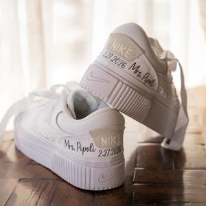 May include: White Nike sneakers with platform soles. The shoes have the Nike logo and the text "Mrs. Pipoli 2.27.2026" printed on the back. The sneakers are on a wooden surface.