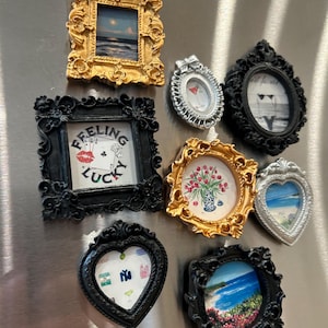 Puede incluir: Una colección de marcos de fotos decorativos en miniatura de varias formas y colores. Los marcos son dorados, plateados y negros, con detalles ornamentados. Algunos marcos contienen pequeñas imágenes de paisajes y otros diseños. Un marco tiene el texto "Feeling Lucky".