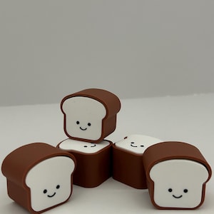 May include: A collection of brown and white bread-shaped erasers. Each eraser has a smiling face on the white bread portion. The erasers are arranged in a playful, scattered composition against a plain background.
