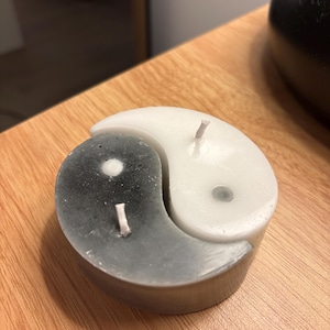 Puede incluir: Una vela con forma de Yin y Yang. La vela está dividida en dos secciones, una blanca y otra gris. Cada sección tiene una mecha. La vela está sobre una superficie de madera.