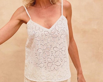 May include: A white camisole top with a V-neck and spaghetti straps. The top is made of a floral eyelet fabric. The floral pattern is made of small, evenly spaced holes. The background is a solid, light tan color.