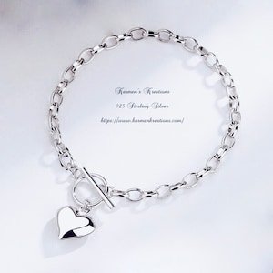 May include: A 925 sterling silver chain bracelet featuring a toggle clasp and a polished heart-shaped charm. The text Karmen's Kreations, 925 Sterling Silver, and https://www.karmenkreations.com/ is printed in the centre of the circular chain.