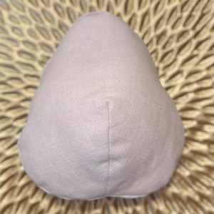 May include: A light purple, teardrop-shaped fabric object. The object is made of a soft, textured material and has a vertical seam down the center. The background is a woven, light brown surface.