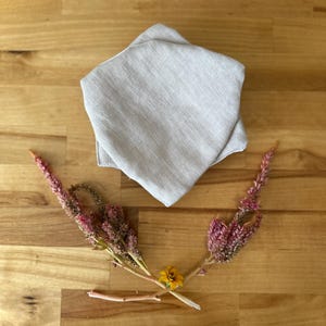 May include: A light beige linen fabric pouch folded into a star shape. The pouch is lying on a wooden surface with a small sprig of pink flowers and a yellow flower.