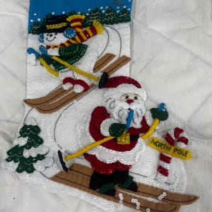 May include: A white felt Christmas stocking featuring a sequined scene of a snowman and Santa Claus skiing. The design includes green pine trees, a blue sky, and a yellow signpost with the text NORTH POLE next to a red and white candy cane.